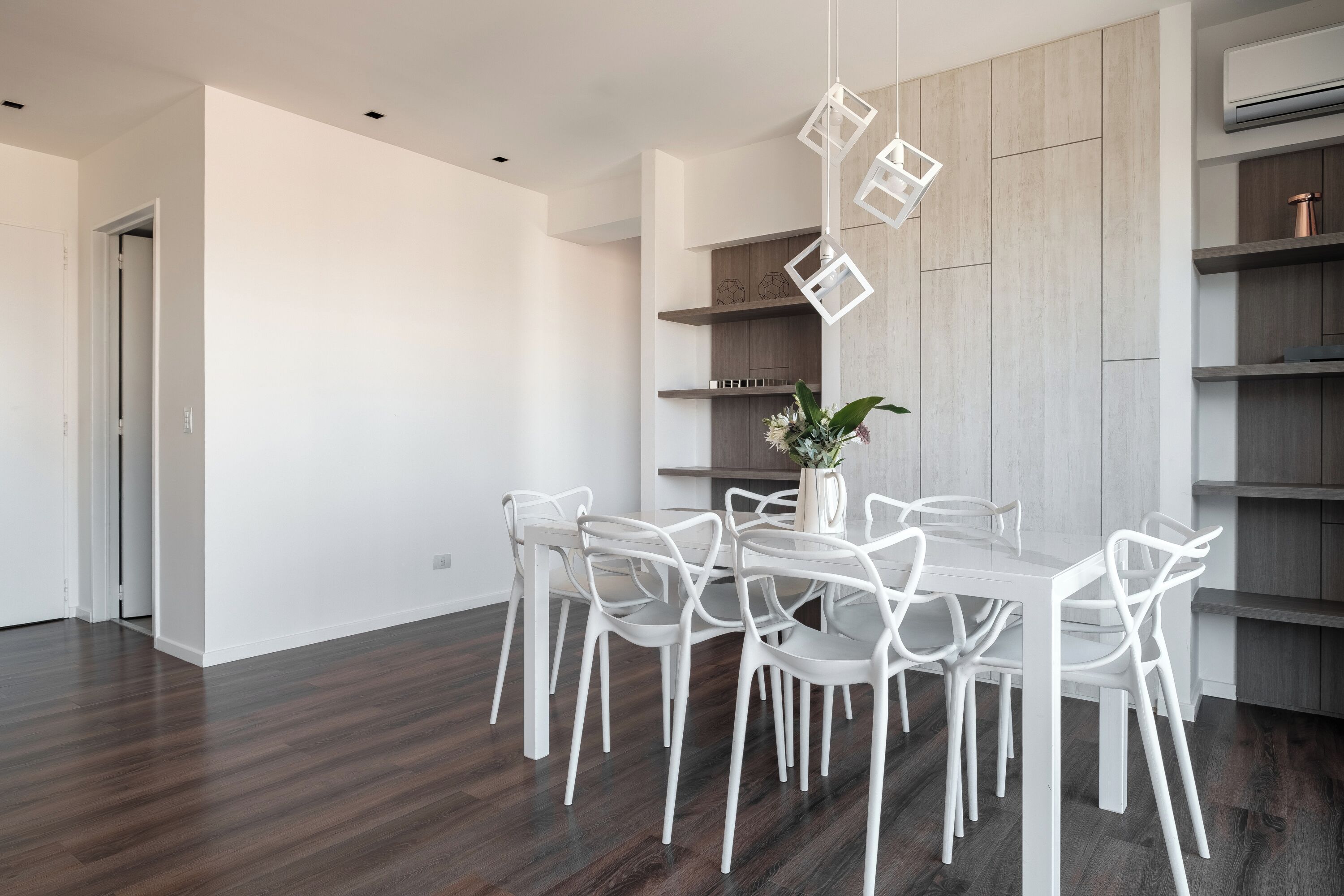 Un'area pranzo moderna con un tavolo rettangolare e sedie bianche, un lampadario a sospensione e scaffalature a parete con una pianta sul tavolo.