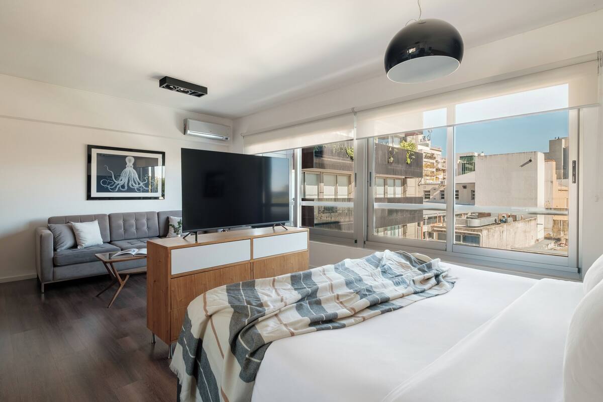 A modern hotel room with a large bed, a flat-screen TV, a sofa, a wooden dresser, and a large window offering a city view.