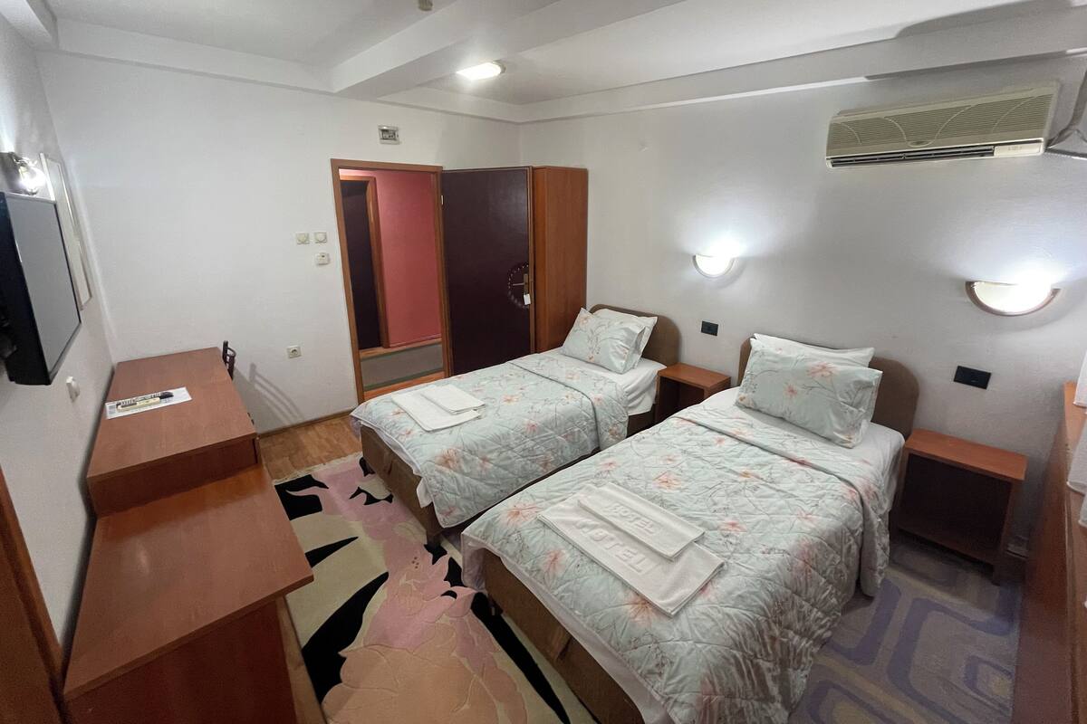 Quarto de hotel com duas camas, uma escrivaninha, um espelho e um ventilador de teto.