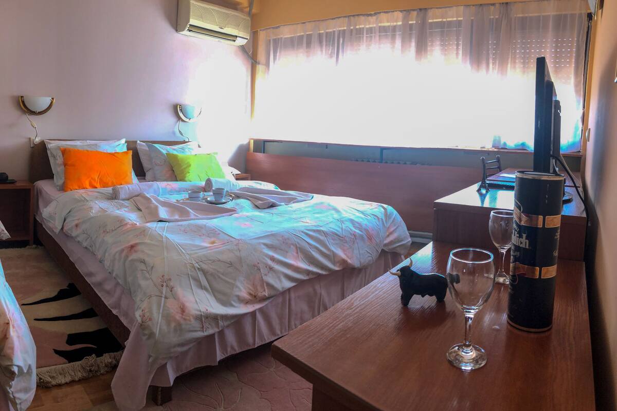 Quarto de hotel com duas camas, uma mesa com computador, um copo de água e uma janela com cortinas.