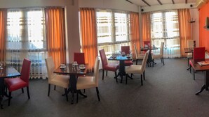 A hotel dining area with multiple tables and chairs, large windows with curtains, and a flat-screen TV mounted on the wall. - Hotel Square (Skopje)