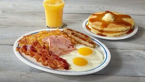 A breakfast plate with bacon, eggs, and sausages, a glass of orange juice, and a stack of pancakes with syrup and butter on a wooden table. - Hotel Square (Skopje)