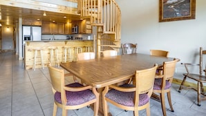A wooden dining area with a large table and chairs, a spiral staircase, and a kitchen in the background.