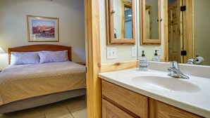 A bedroom with a bed, a wooden headboard, a large mirror, a bathroom with a sink and mirror, and a framed picture on the wall.