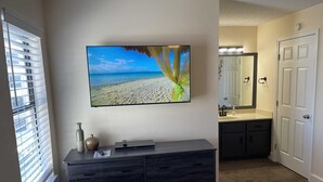 Une chambre d’hôtel dotée d’un grand téléviseur à écran plat fixé au mur, d’une commode en bois foncé, d’une salle de bain avec un lavabo et un miroir, et d’une fenêtre avec des stores.