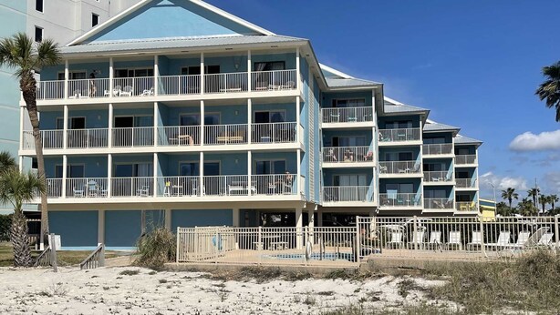 Un hôtel à plusieurs étages doté de balcons, situé près d’une plage de sable fin, où la végétation est clairsemée.