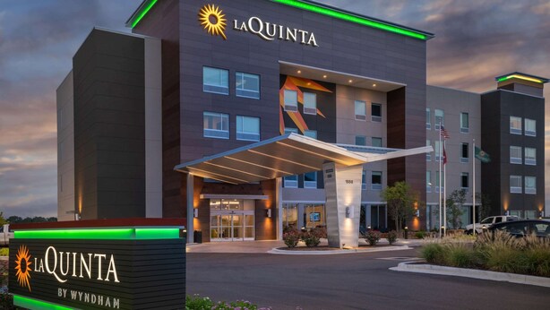 Ein modernes Hotel mit markantem Eingang, einem Schild mit der Aufschrift La Quinta by Wyndham und freier Sicht auf den Parkplatz.