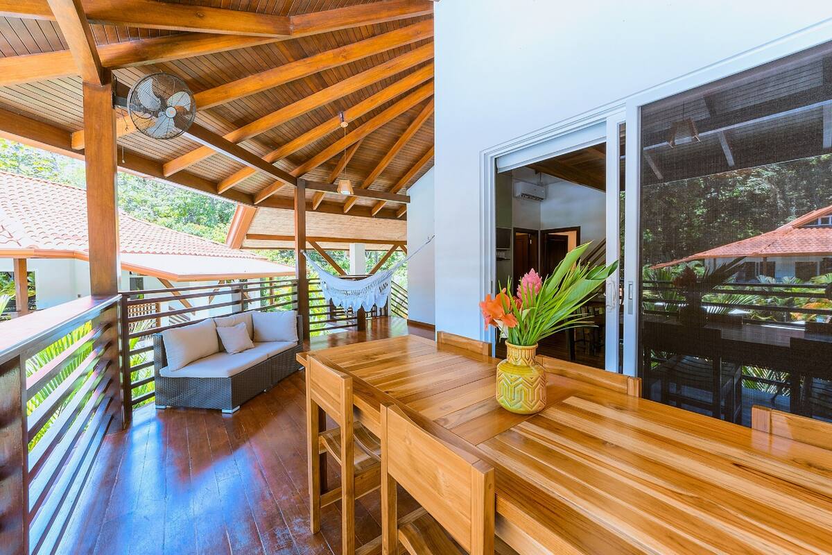 Une terrasse en bois avec un hamac, une table en bois ornée d’un vase de fleurs et un canapé en osier.
