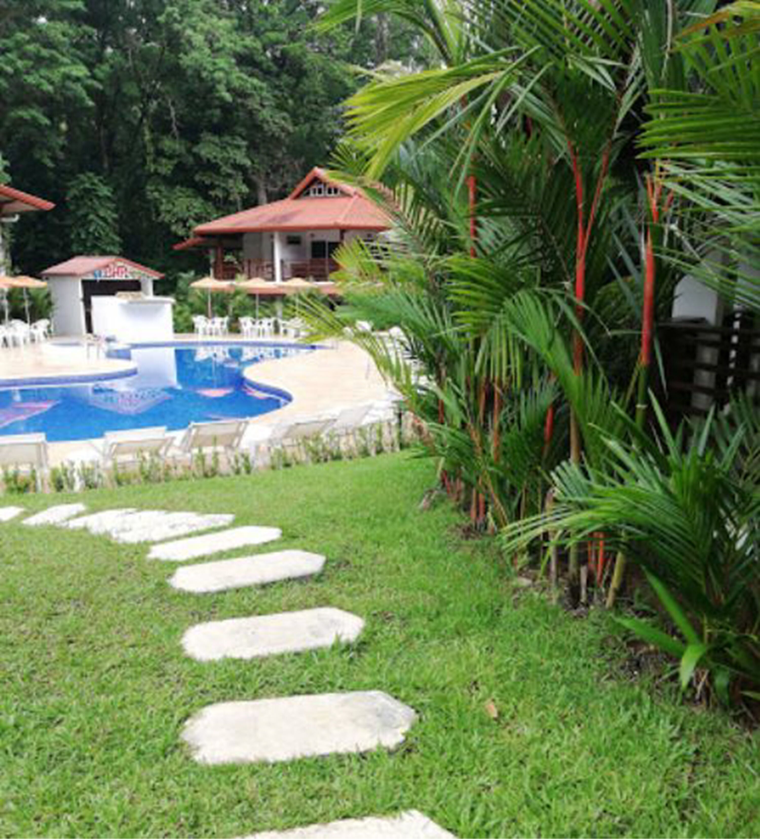 A pool area with stepping stones leading to a lounge area, surrounded by palm trees and a building with a red roof.
