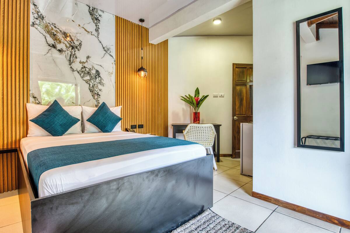 Une chambre à coucher moderne avec un grand lit, un lambris en bois, un mur en marbre et un petit bureau agrémenté d’une plante.