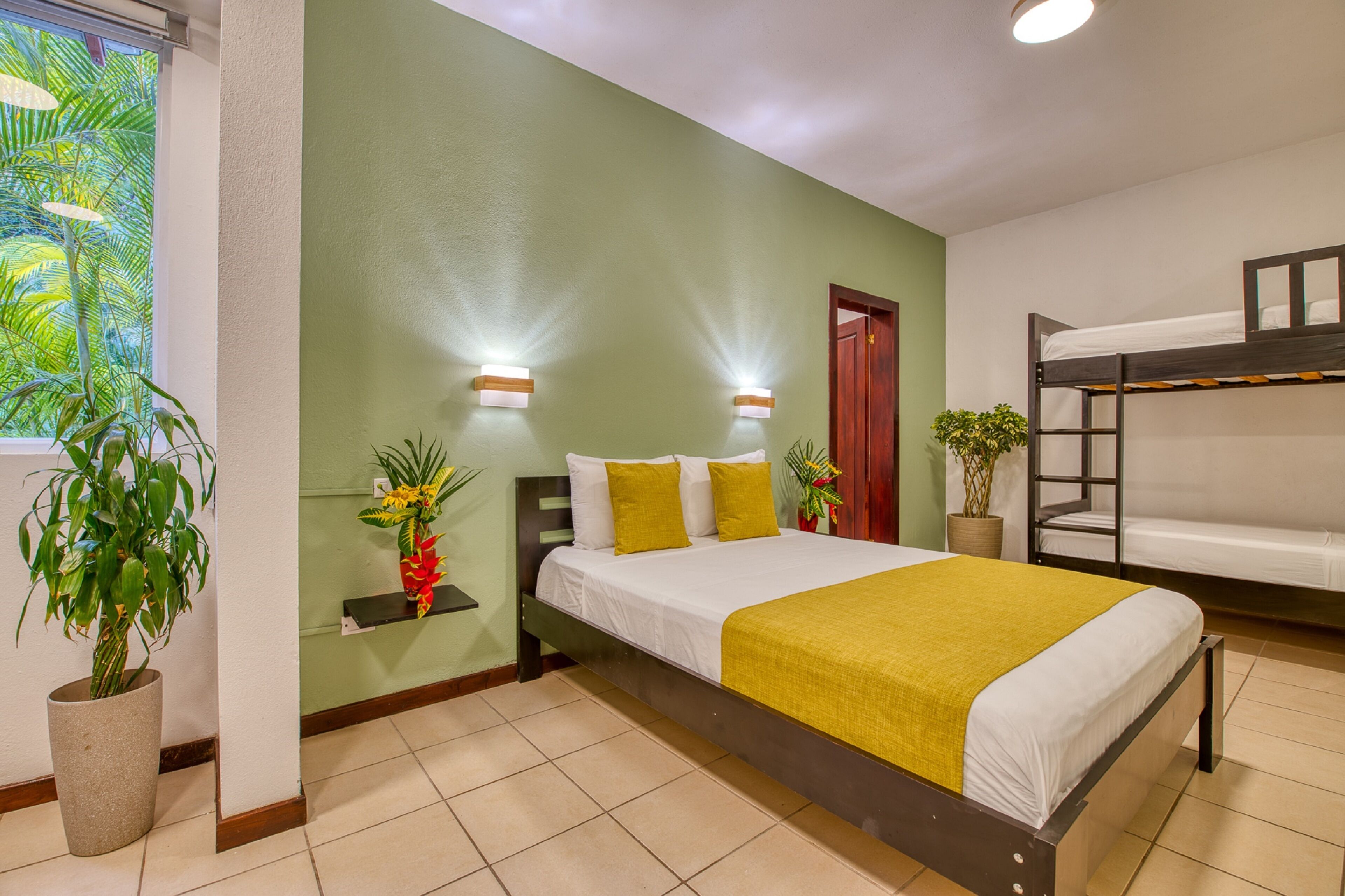 A hotel room with a bunk bed, a large bed with a yellow blanket, two wall-mounted lights, a potted plant, and a window with a view of palm trees.
