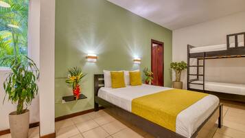 A hotel room with a bunk bed, a large bed with a yellow blanket, two wall-mounted lights, a potted plant, and a window with a view of palm trees.