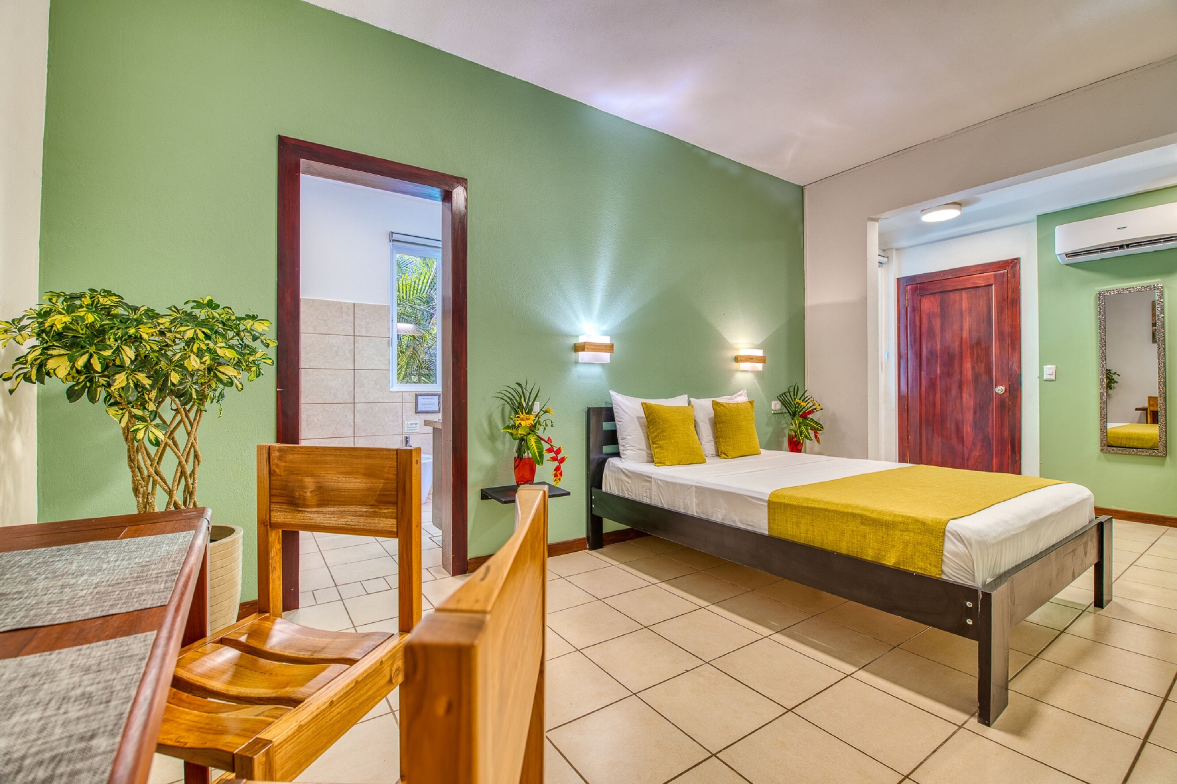 A hotel room with a bed, a wooden chair, a small table, and two potted plants.