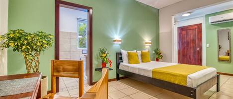 Habitación de hotel con una cama, una silla de madera, una mesita pequeña y dos plantas en macetas.