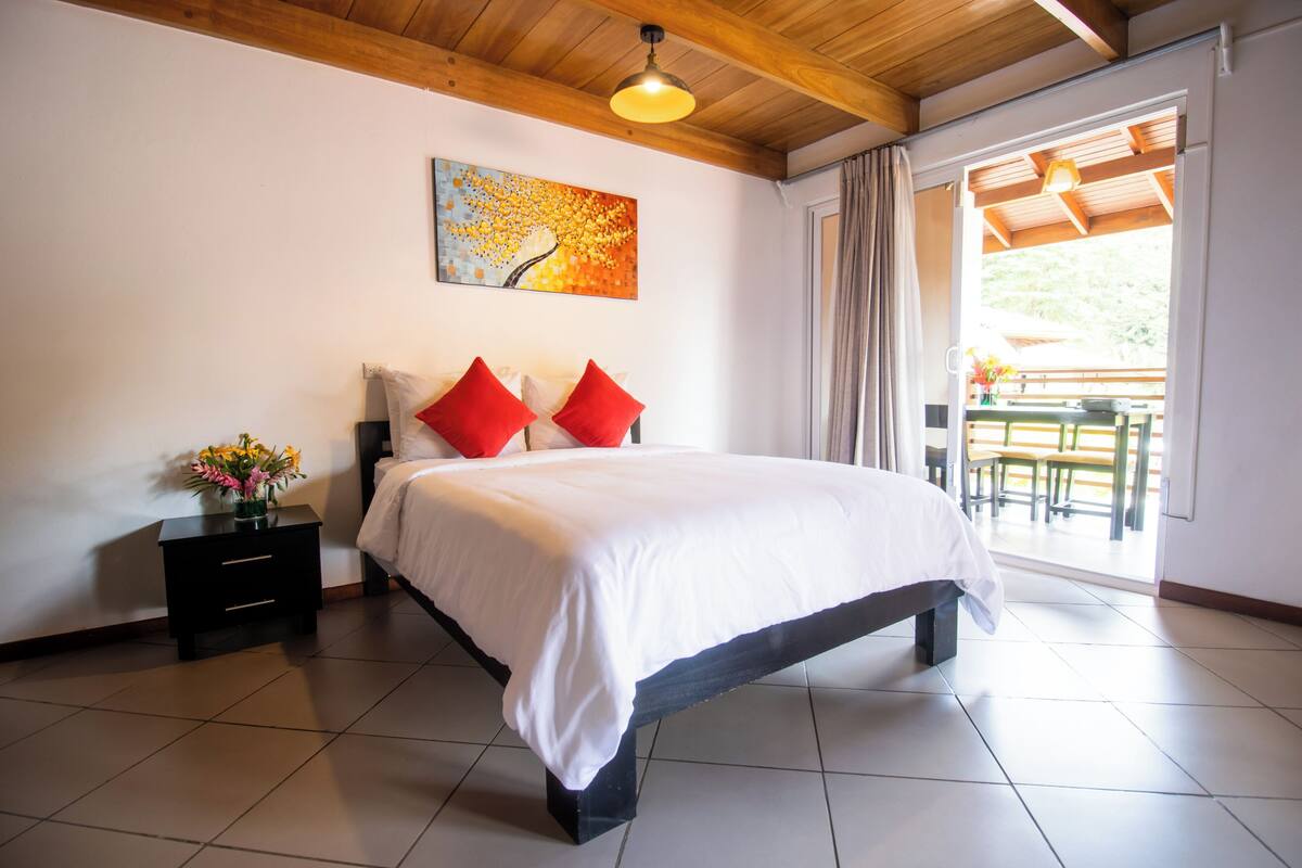Une chambre à coucher avec un lit, une table de chevet ornée d’un vase de fleurs, un tableau accroché au mur et une vue sur une terrasse extérieure.