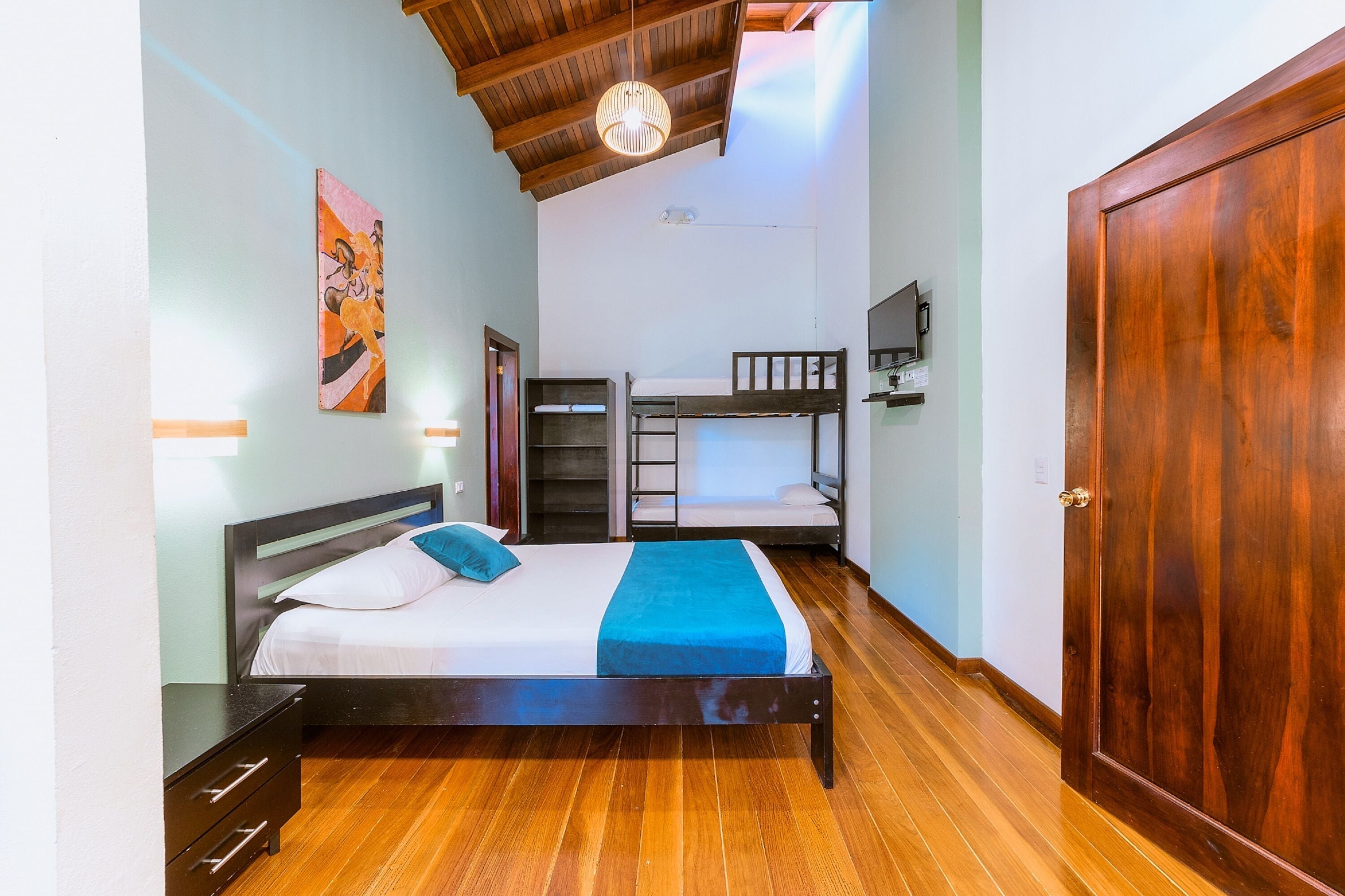 A bedroom with a bunk bed, a wooden floor, a bed with a blue blanket, a nightstand, a wall-mounted TV, and a wooden door.
