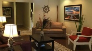 A living room with a sofa, a coffee table, a red chair, a lamp, and a ceiling fan. - Admiral's Club, wide cove, spa, pool/ping pong/air hockey/poker tables (Gainesville)
