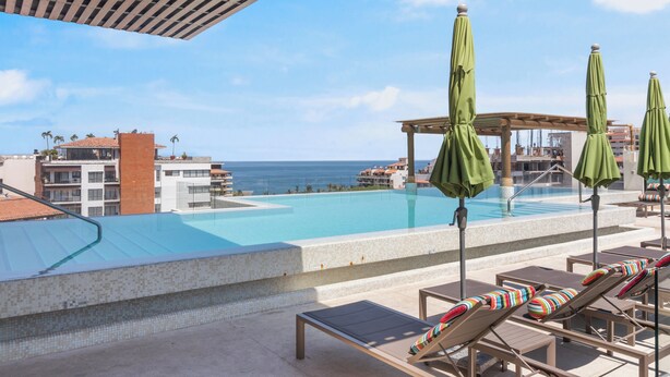 Un área de piscina en la azotea con sillones de descanso y sombrillas, con vistas a un cielo azul despejado y una lejana vista del océano.