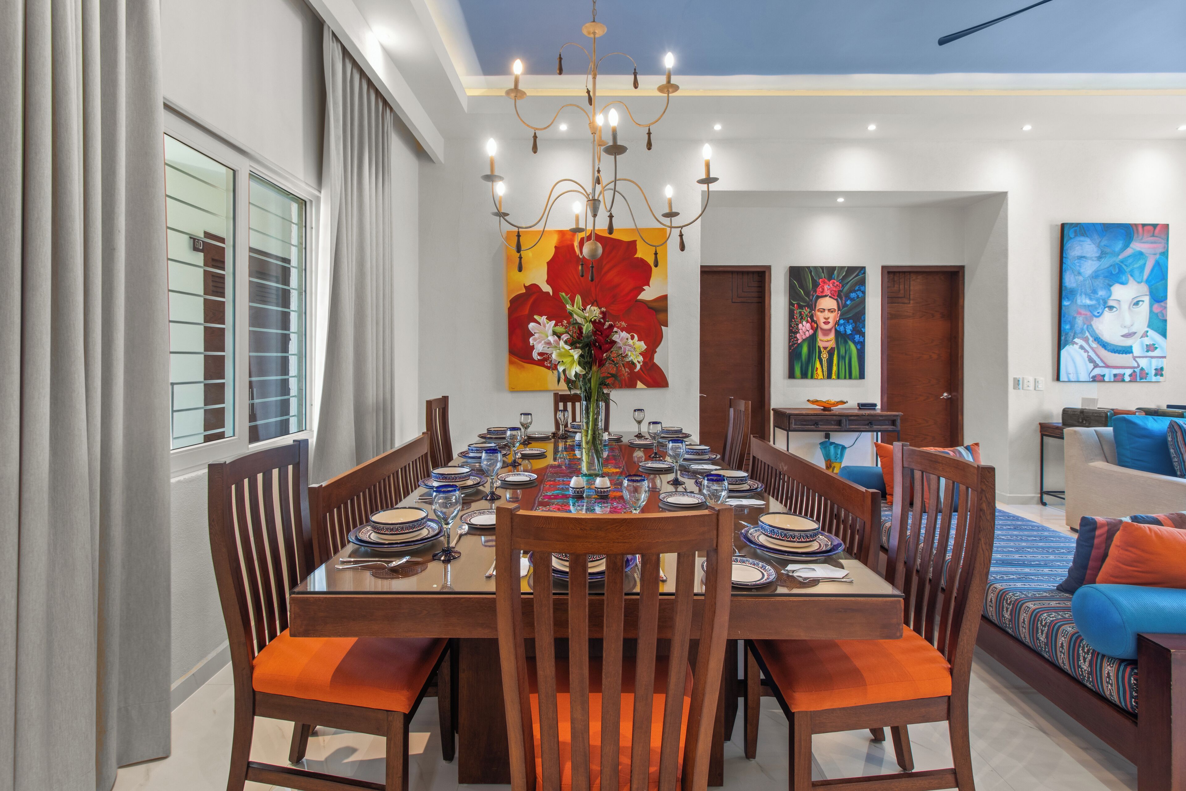 A dining area with a large table set for six, surrounded by wooden chairs with orange cushions. A chandelier hangs above the table, and a large floral painting is visible on the wall behind the table.