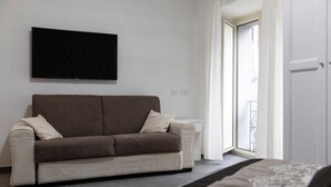 A modern living room with a sofa, a flat-screen TV mounted on the wall, and a large window providing natural light. - Alambrado Rooms & Suites (Naples)