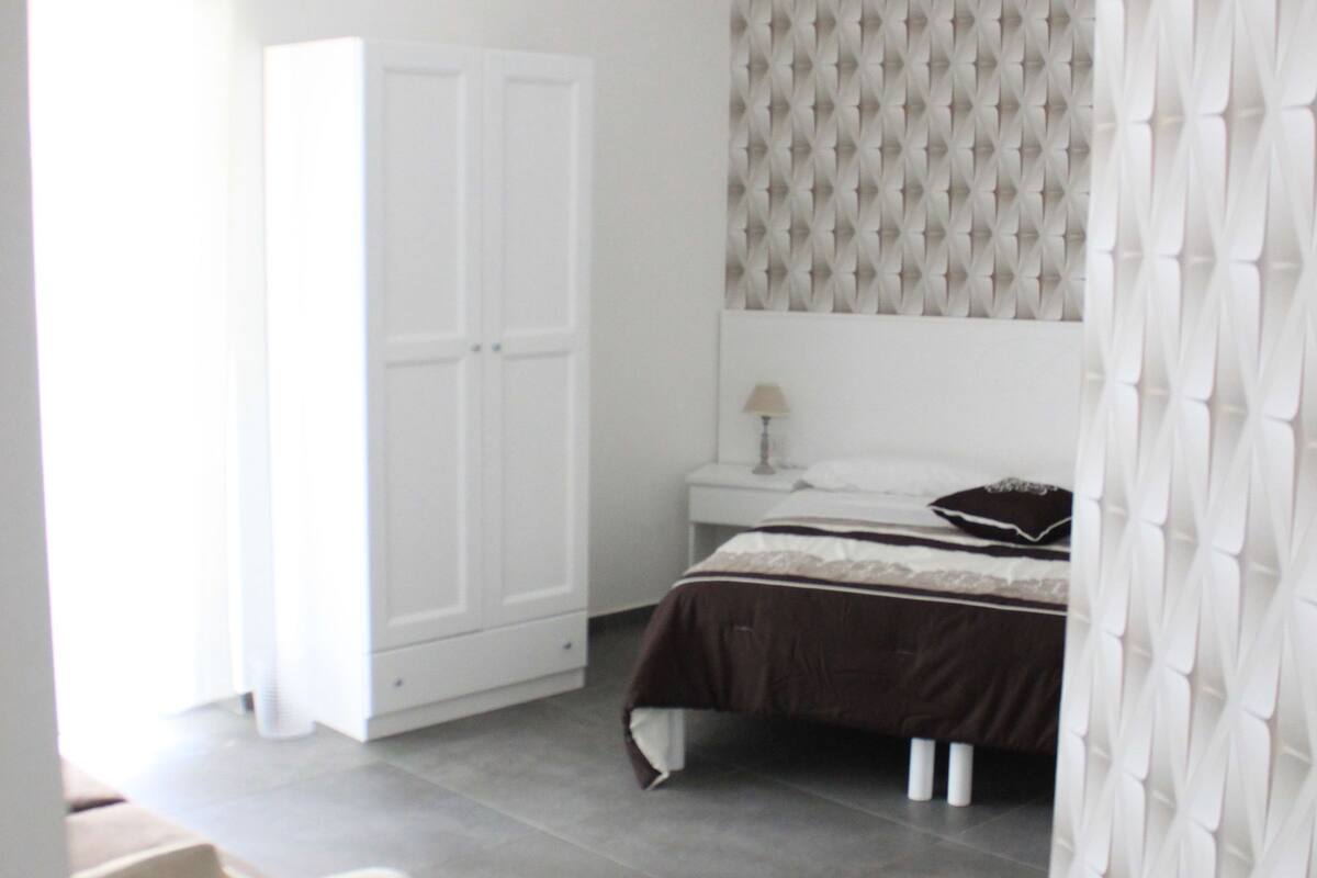 A bedroom with a bed, a wardrobe, a bedside table, and a window with curtains.