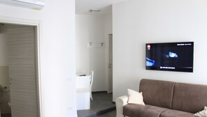 A modern hotel room with a brown sofa, a flat-screen TV mounted on the wall, and a white door leading to another room. - Alambrado Rooms & Suites (Naples)