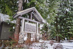 A snowy Greek-themed sign with a stone base and a wooden roof, surrounded by snow-covered trees and a wooden building in the background.