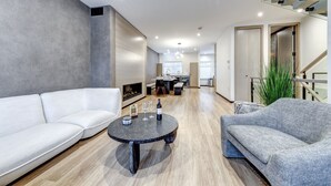 Een moderne woonkamer met een witte bank, een grijze fauteuil en een ronde salontafel met wijnglazen en een fles wijn.