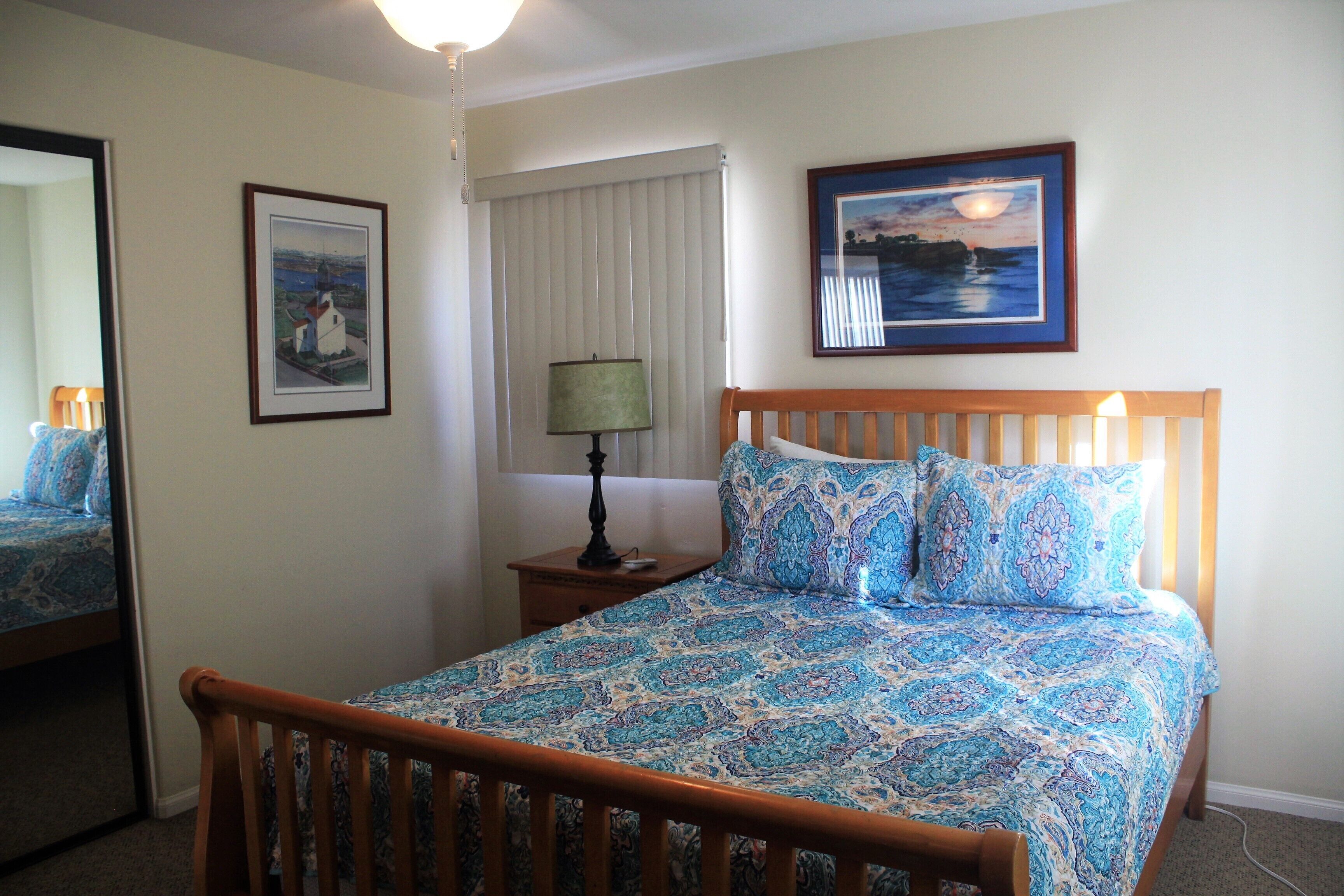 A bedroom with a wooden bed, a nightstand with a lamp, a window with blinds, and two framed pictures on the wall.