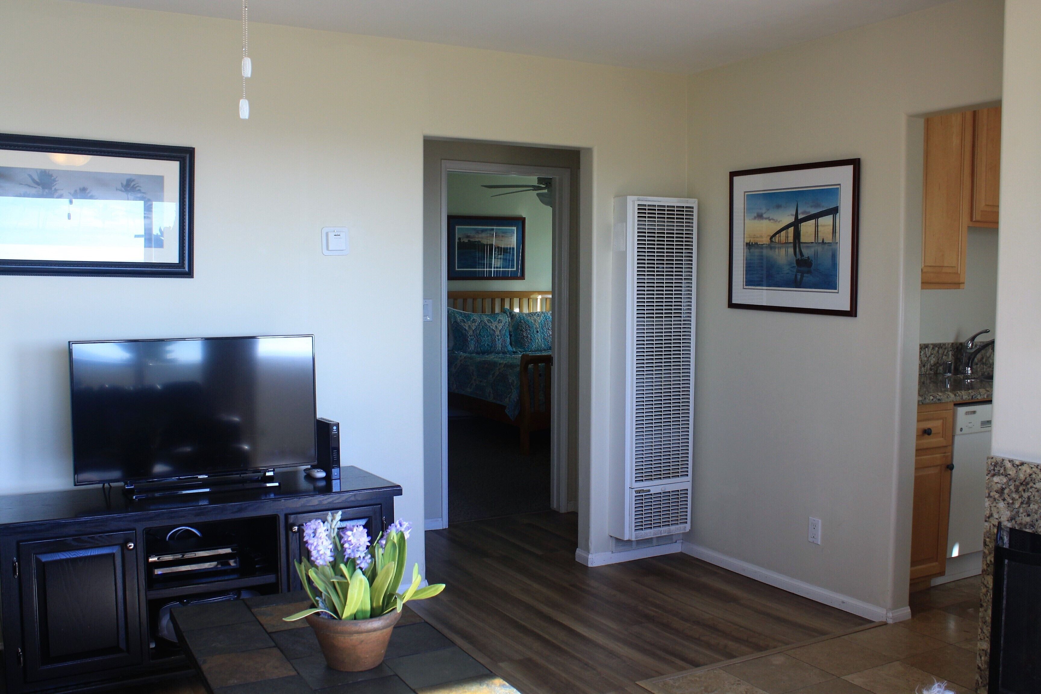 A living room with a flat-screen TV, a dark wooden entertainment unit, a potted plant, and a hallway leading to a bedroom.