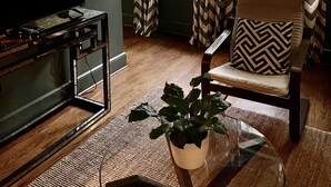 Ein Wohnzimmer mit Flachbildfernseher, einem Holztisch mit Pflanze, einem gepolsterten Sessel und einem Fenster mit gemusterten Vorhängen.