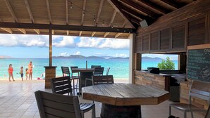 A beachside restaurant with wooden tables and chairs, a view of the ocean, and a chalkboard menu listing food items. - Gorgeous boho at the beach walkout oceanfront condo in Sapphire D building. (St Thomas)