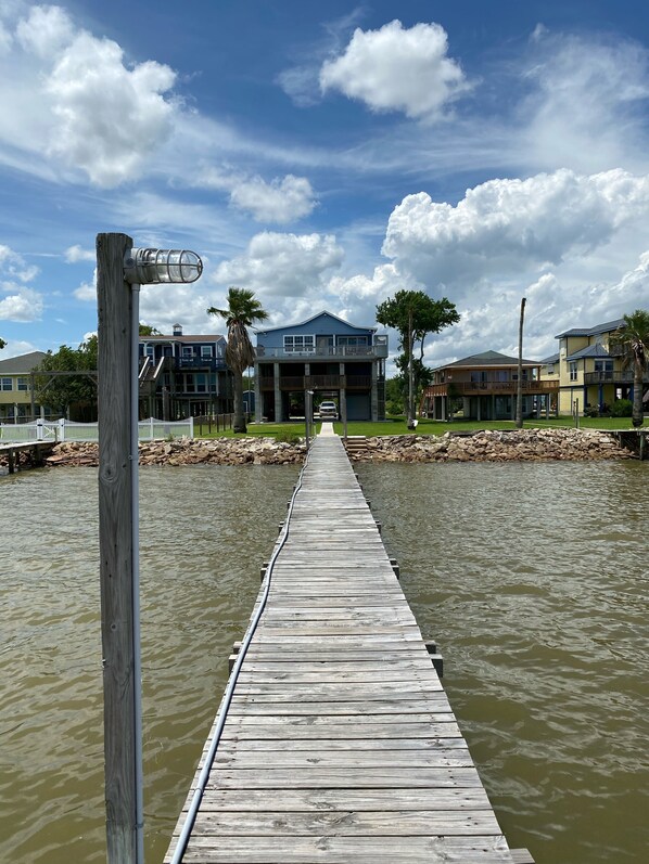 A wooden pier leading to a house with a balcony, surrounded by water and palm trees.