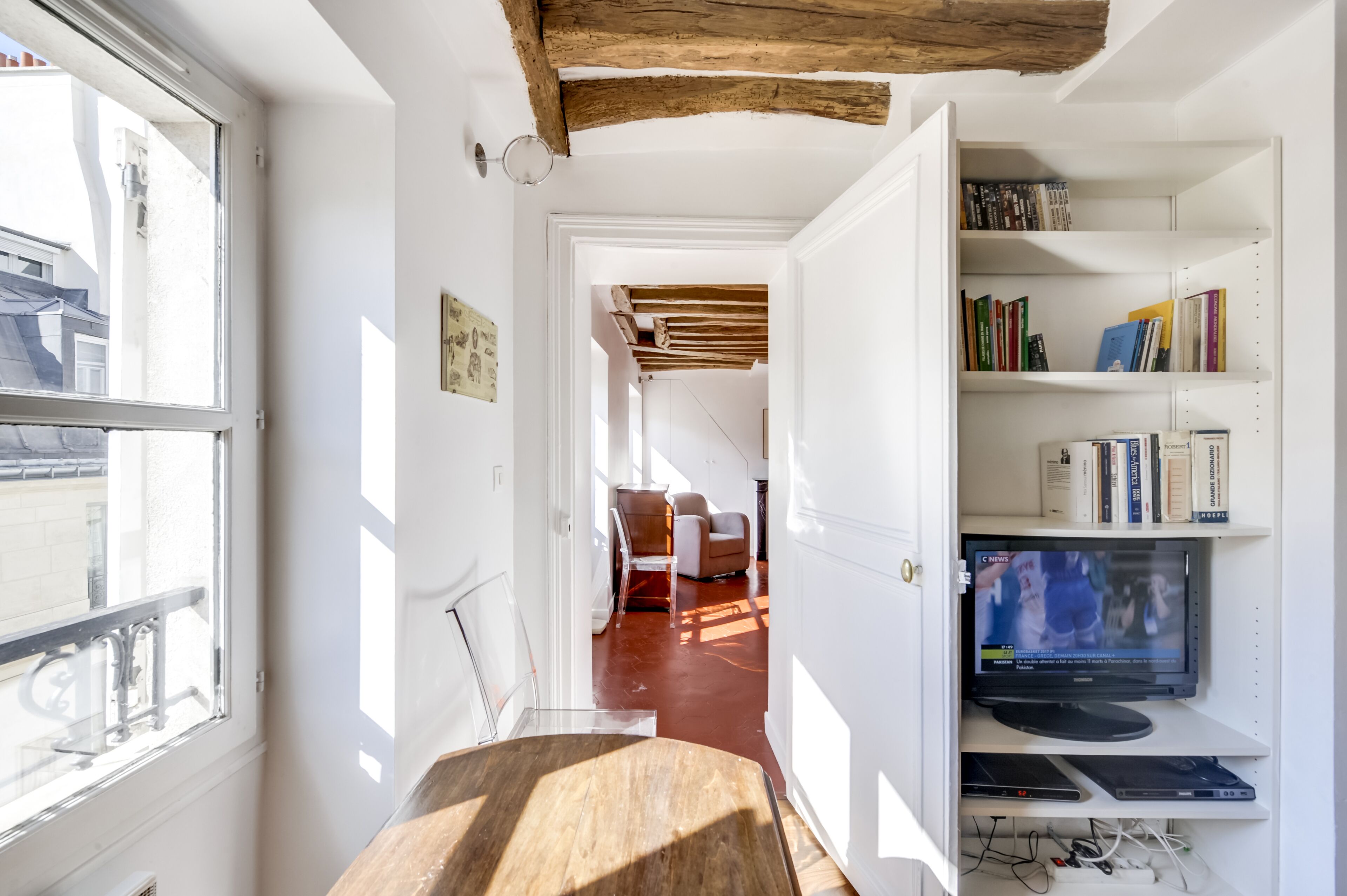 Een kamer met een houten plafond, een boekenplank vol boeken, een televisie en een glazen schuifdeur naar een andere kamer.
