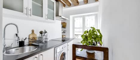 A modern kitchen with white cabinets, a stainless steel sink, a built-in washing machine, and a wooden ceiling beam.