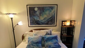 A bedroom with a bed featuring a blue and white abstract design, a night sky painting above the bed, a bedside lamp, and a nightstand with a clock.