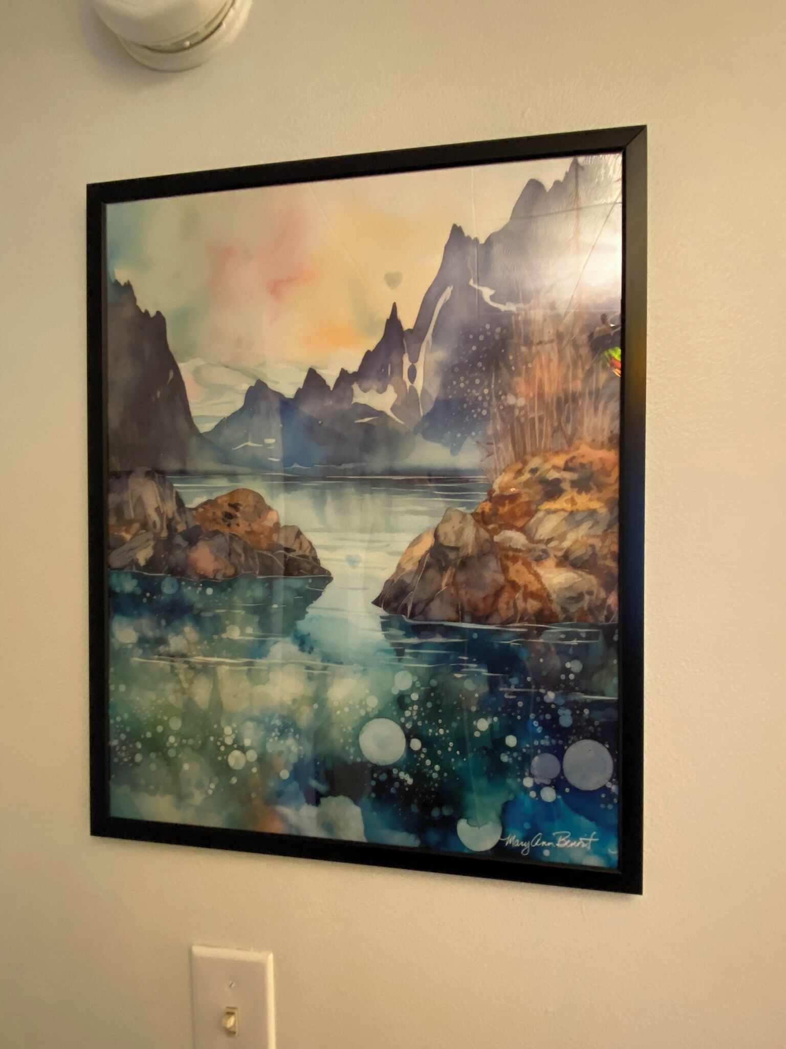 Un tableau encadré représentant un paysage montagneux avec un lac, accroché à un mur uni.