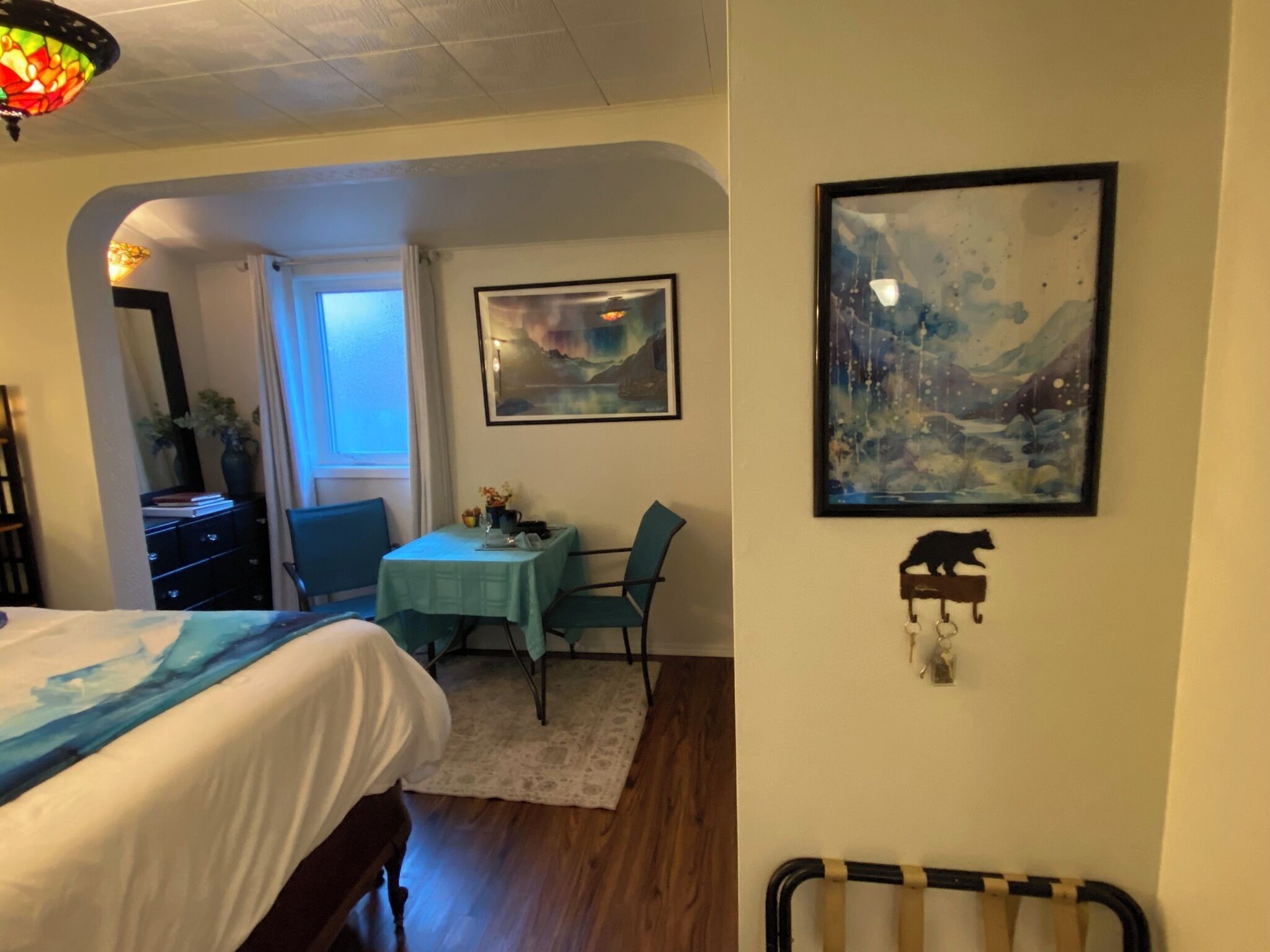 A room with a bed, a dining table with chairs, a painting, and a small shelf with a cat figurine.