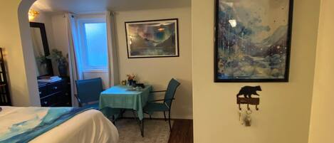 A room with a bed, a dining table with chairs, a painting, and a small shelf with a cat figurine.