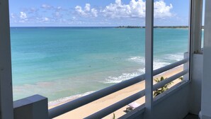 Blick vom Balkon auf einen Strand mit klarem türkisblauem Wasser und einer in der Ferne liegenden Insel.