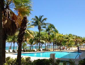 A pool area with lounge chairs and palm trees, overlooking a body of water.