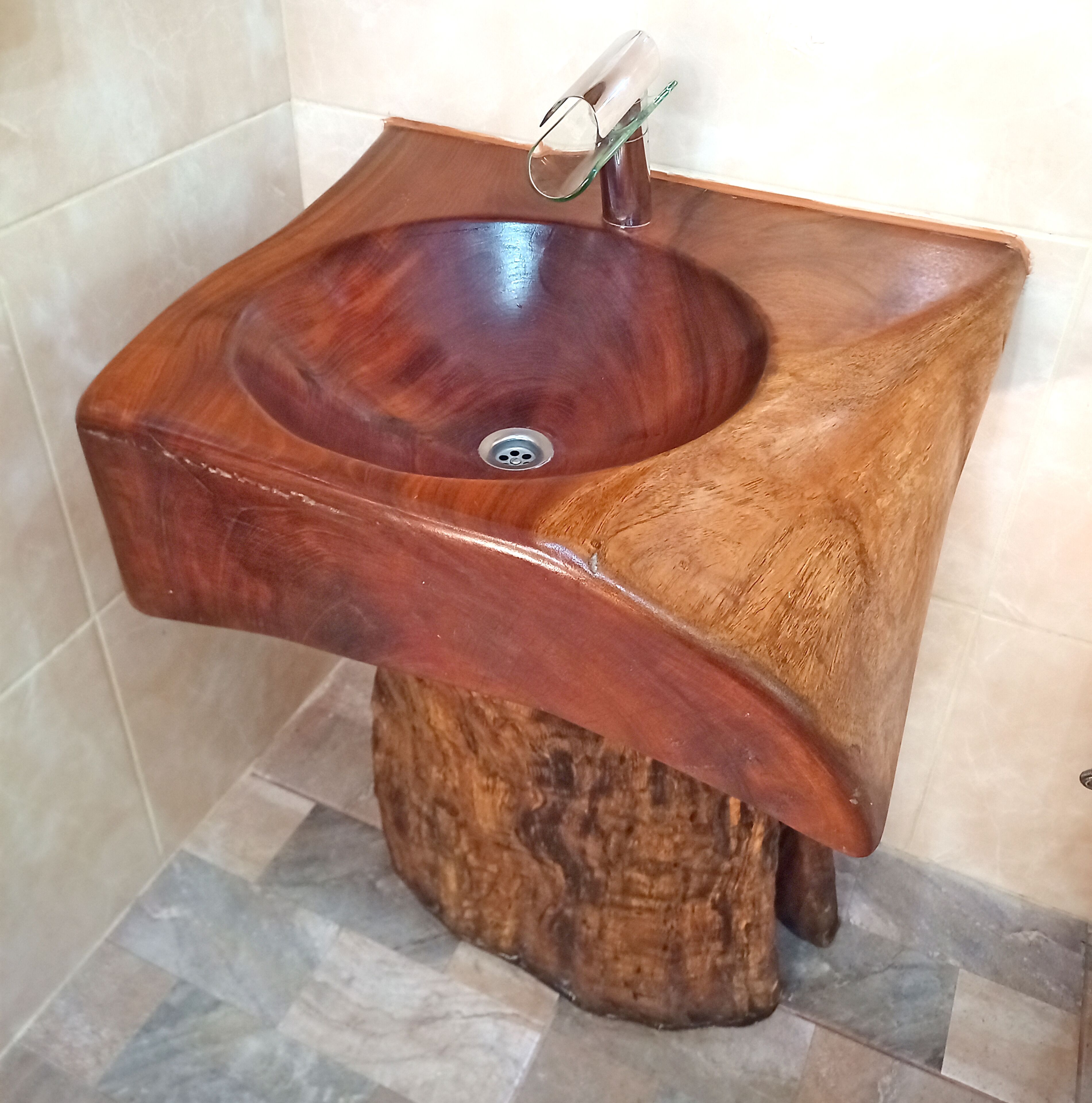 Un évier en bois au design original, installé sur un socle en forme de tronc d’arbre, dans une salle de bain dont le sol est recouvert de carreaux.
