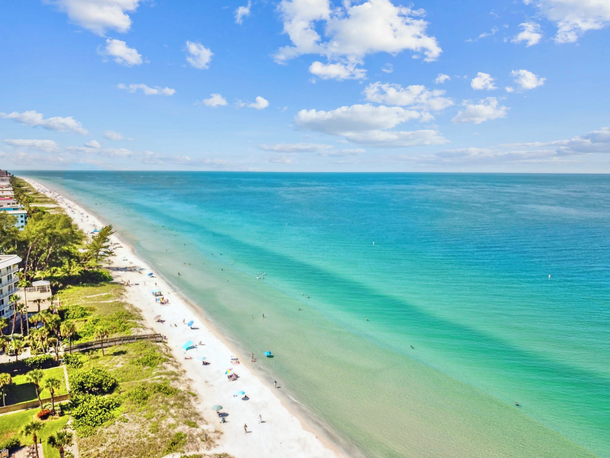 Une plage aux eaux turquoise et limpides, un rivage sablonneux et une rangée de bâtiments le long de la côte.