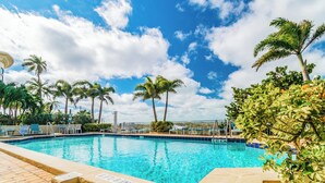 A swimming pool with clear blue water, surrounded by palm trees and lounge chairs, under a partly cloudy sky. - 2 Bedroom In Indian Rocks Beach, FL - Gulf Side Complex w/ Direct Beach Access (Indian Rocks Beach)