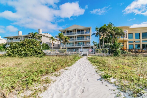 A beachfront hotel with a sandy pathway leading to the entrance, surrounded by palm trees and other greenery.