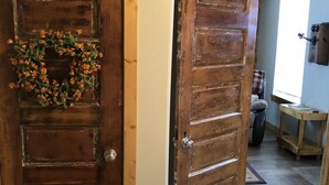 Uma porta de madeira com uma coroa, uma cama com colcha estampada e um corredor que leva a um quarto com janela e prateleira.
