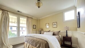 Une chambre à coucher avec un grand lit, une table de chevet, une lampe et deux fenêtres avec des rideaux.