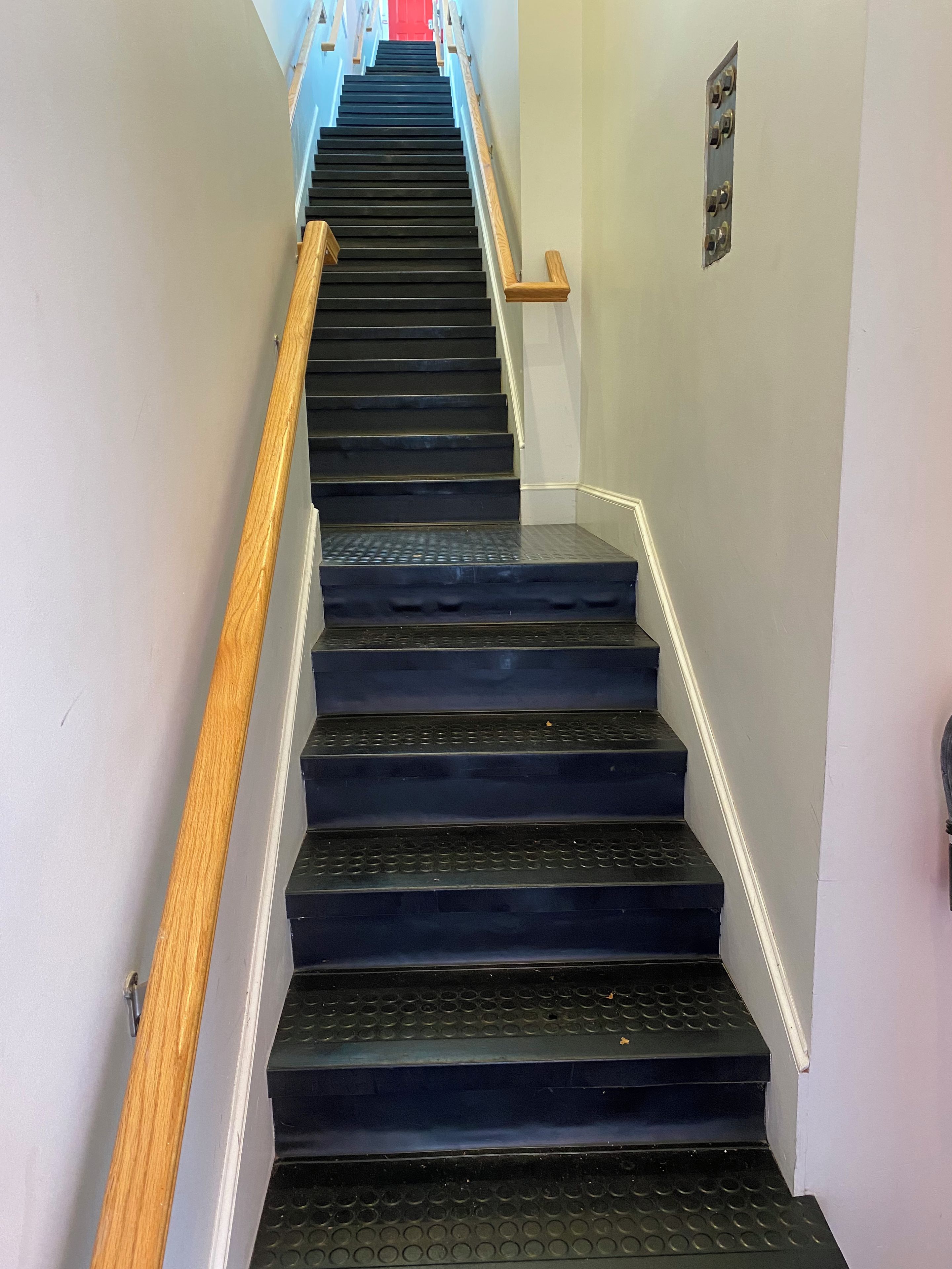 A staircase with a wooden handrail and a metal banister, leading to a red door at the top.