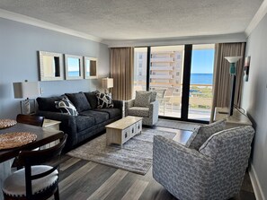 A modern living room with a sofa, armchair, and dining table, large windows with a view of the ocean, and a balcony with outdoor furniture. - Oceanfront 6th Floor- A Must See Remodel! Farmhouse Decor + Beach Locker! (Myrtle Beach)