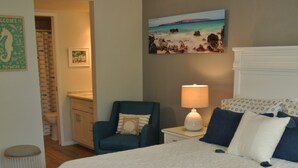 Una camera da letto con un letto grande, una sedia, un comodino con una lampada, un quadro appeso e un bagno visibile attraverso una porta aperta.